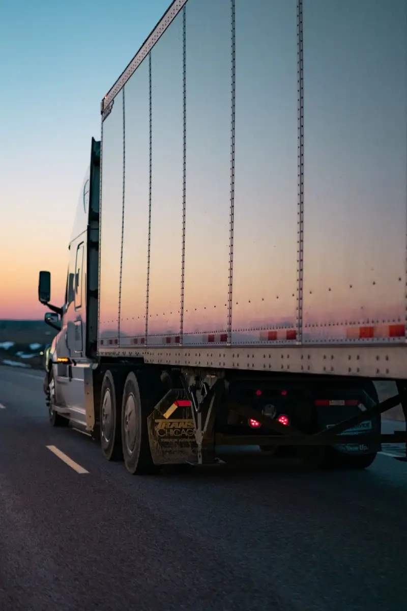 A semi-truck driving at sunset with visible mud flaps installed behind the rear wheels to protect the vehicle and others from road debris.
