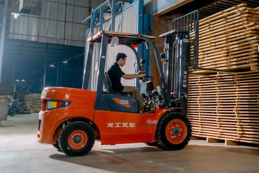 Forklift operator driving an orange forklift past tall stacks of wooden planks in a warehouse